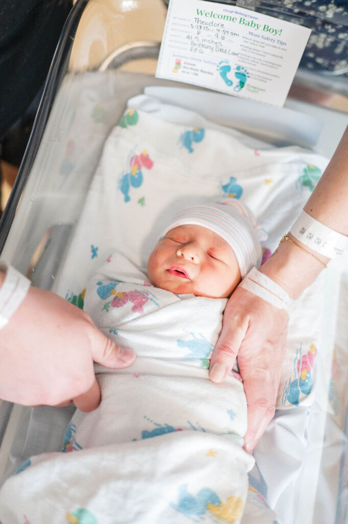 Newborn baby swaddled in hospital blanket with mom and dad's hands on him at Clough Birthing Center
