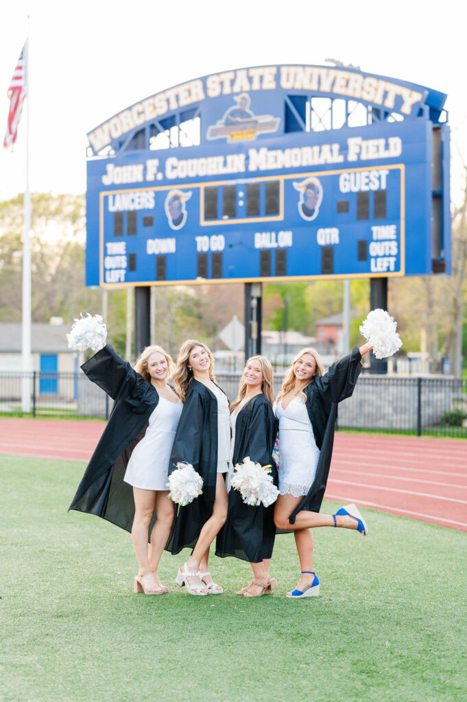 Four grads on the field in cap and gown with pom poms