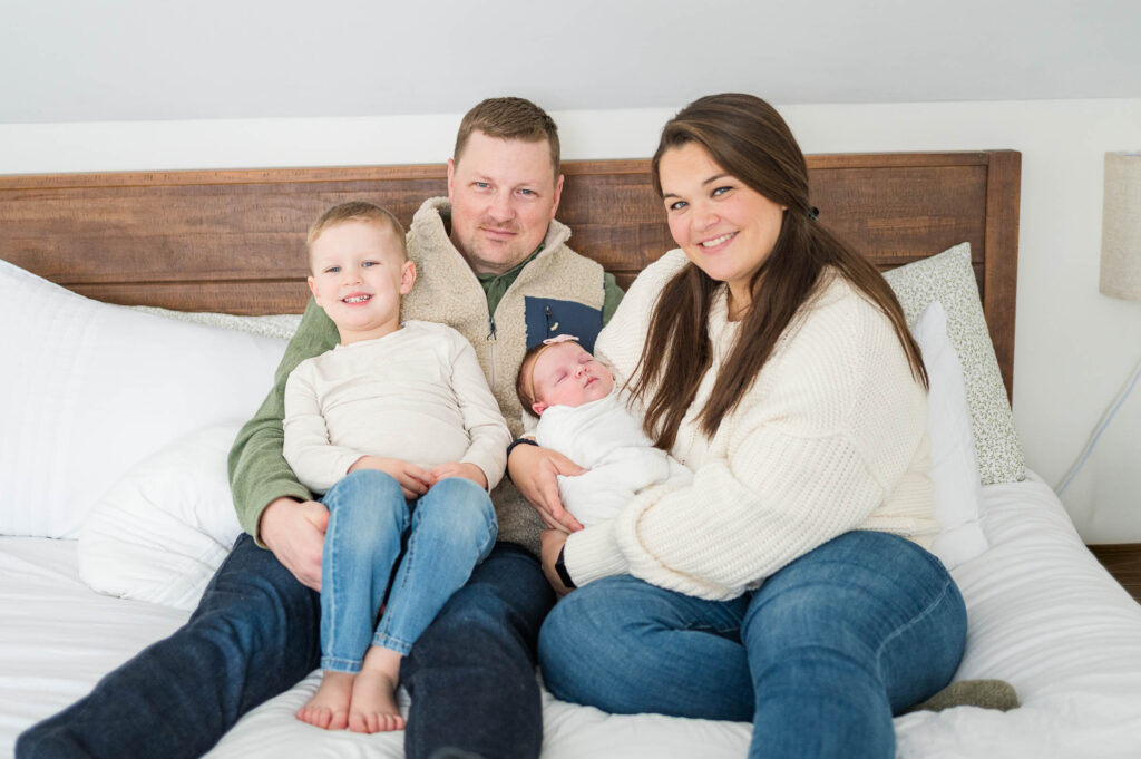 Walpole, MA newborn photographer's image of a family sitting on the bed with little boy and newborn girl smiling at camera
