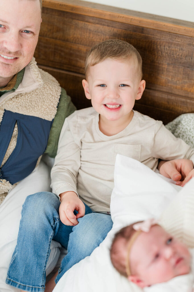 Focus on little boy smiling at camera with dad and newborn sister in photo in Walpole, MA