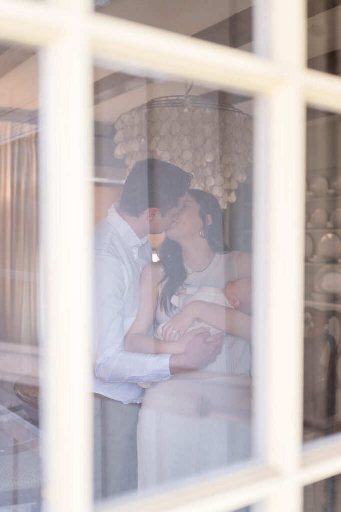Image of mom and dad kissing with newborn baby in arms taken through window